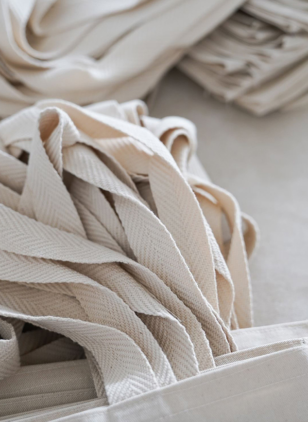 Fitas de tecido em tom cru com textura natural, empilhadas e dobradas, usadas na produção de brindes personalizados